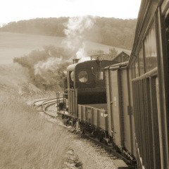 Eine kleine Dampflok in hügeligem Gelände vom fahrenden Zug aus gesehen.