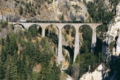 Ein Personenzug mit Dampflok überquert einen steinernen Viadukt in den Alpen.