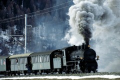 Stark qualmende Dampflokomotive vor einem Museumszug in verschneiter Landschaft.