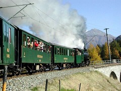 Zug mit Schlepptender–Lokomotive und grünen Wagen dampft eine Steigung hinauf, schräg von hinten gesehen.