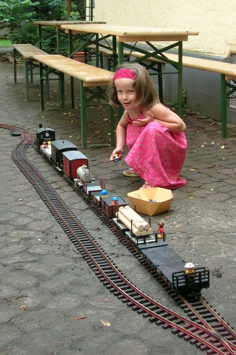 Ein kleines Mädchen hockt im Hof vor einem Zug auf den Gleisen und spielt mit der Eisenbahn und Figuren.