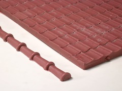 Ein rotbraunes Stück Dachplatte mit Formziegeln und eine Stange Firstziegel, beide nur teilweise sichtbar.