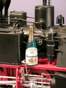 Eine Champagner–Flasche steht auf dem Umlauf einer Modell–Dampflok.