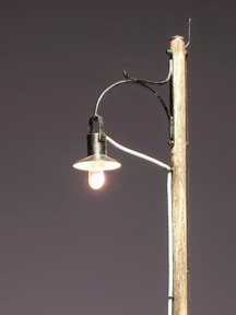 Modell: das Oberteil einer Straßenlampe mit Holzmast und brennender Glühlampe.