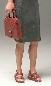 Der untere Teil einer stehenden Frauenfigur mit Rock, Ledertasche und Sandalen.