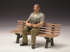 Figur: Mann im kurzärmeligen Hemd auf einer Bank, eine Tasche auf dem Schoß.