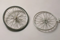 Ein Modellfahrrad–Rad mit vielen dünnen Metallspeichen, daneben eines aus Kunststoff mit wenigen dickeren Speichen.