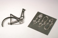 Schwarz gespritzter Fahrrad–Rahmen, die eingesetzte Kette samt Zahnrädern und daneben ein Ätzblech mit Kettenteilen.