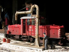 Eine rote Diesellok bei einem größeren Wasserkran, dahinter ein Bahnbeamter.