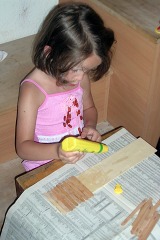 Ein kleines Mädchen im rosa Hemd klebt Eisstäbchen auf ein Stück Sperrholz.