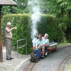 Eine kleine blaue Lok mit einem Sitzwagen fährt durch einen Park.