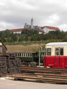 Blick auf Bahnhofsgleise, Triebwagen, Reisezugwagen, auf dem Hügel eine Abtei.