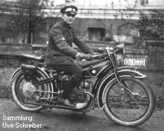 Ein Mann mit einer Schirmmütze und eine Motorradbrille darauf sitzt auf einem Motorrad und blickt gut gelaunt zum Fotografen.