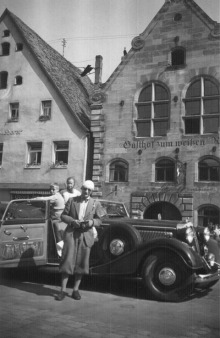 Ein Mann mit Knickerbocker–Hosen vor einem Auto mit zwei Kindern darin.