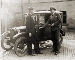 Foto: Zwei Herren im Anzug vor kleiner Limousine, der rechte mit Hut und der Hand am Türgriff.