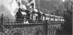 Zwei Schlepptender–Dampflokomotiven ziehen einen Personenzug über einen steinernen Viadukt.