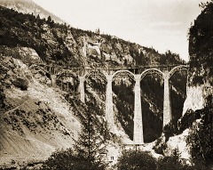 Blick aus einer Schlucht auf einen halbfertigen, steinernen Viadukt mit sehr hohen Stützpfeilern.