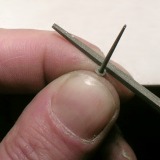 Eine Rundfeile zwischen Daumen und Zeigefinger, darauf ein winziges Metallscheibchen, das mit der Feile entgratet wird.