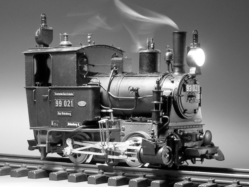 Schwarzweiß–Foto: Leuchtende und rauchende, zweiachsige Modell–Dampflok schräg von vorne.