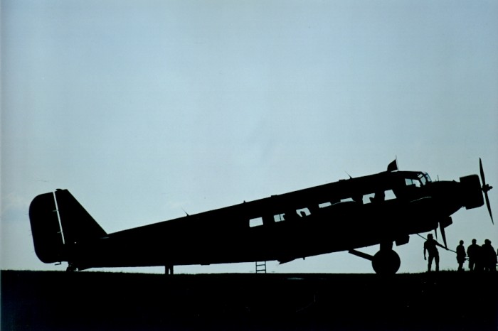 Flugzeug in der Dämmerung als Schattenriß von der Seite. Davor stehen einige Menschen.