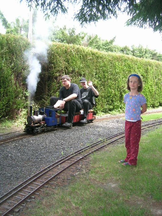 Zwei Männer auf dem Sitzwagen einer Echtdampf–Lok, neben den Gleisen ein Kind.