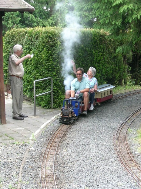 Eine kleine blaue Lok mit einem Sitzwagen fährt durch einen Park.