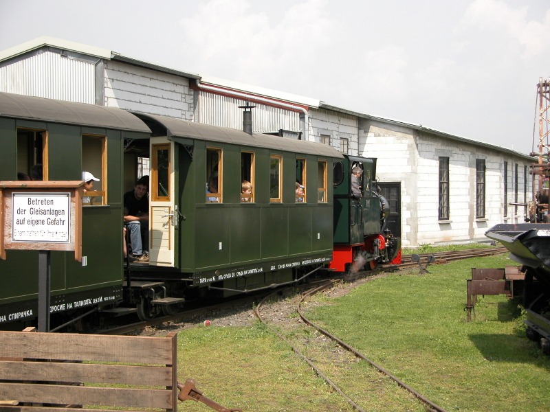 Eine Feldbahn–Dampflok fährt mit Kleinbahn–Reisezugwagen aus dem Bahnhof.