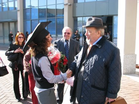 Ein bärtiger Mann mit Hut gratuliert einer Doktorantin auf einem Hof.