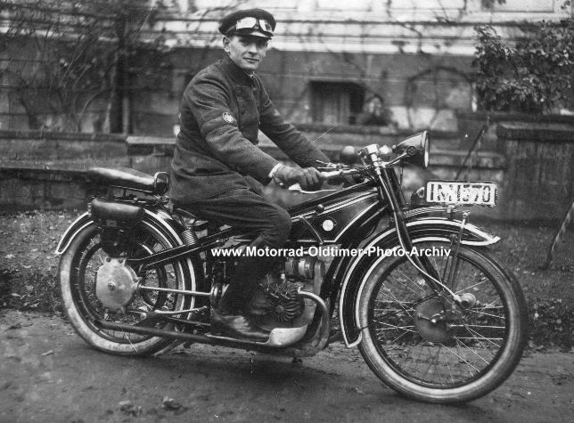 Ein Mann mit einer Schirmmütze und eine Motorradbrille darauf sitzt auf einem Motorrad und blickt gut gelaunt zum Fotografen.