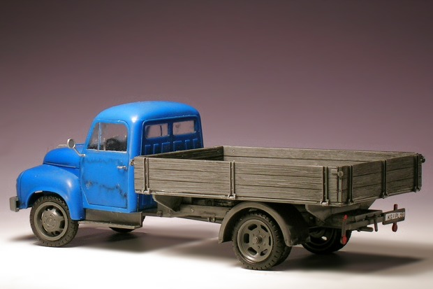 Modellfoto: kleiner Lastwagen mit grauer Prische und blauem Führerhaus, schräg von hinten gesehen.