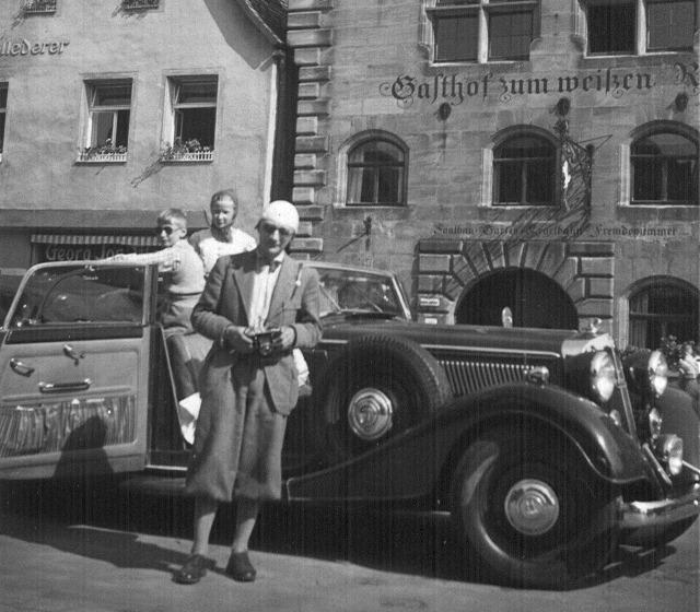 Ein Mann mit Knickerbocker–Hosen vor einem Auto mit zwei Kindern darin.