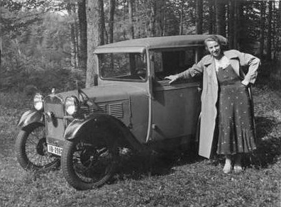 Eine Frau in Kleid und Mantel steht vor dem Kleinwagen beim Waldrand.