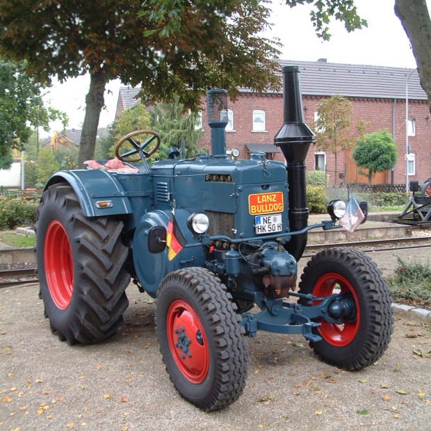 Blick von schräg vorne auf einen blauen Traktor mit roten Felgen und hinteren Kotflügeln, der Führerstand ist offen.