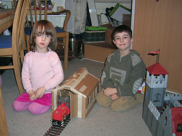 Foto: Zwei Kinder mit aus einem Karton gebastelten Lokschuppen, darin eine kleine rote Diesellok.