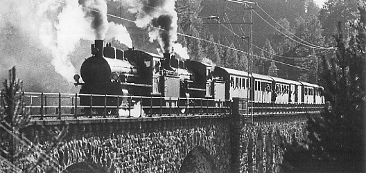 Zwei Schlepptender–Dampflokomotiven ziehen einen Personenzug über einen steinernen Viadukt.