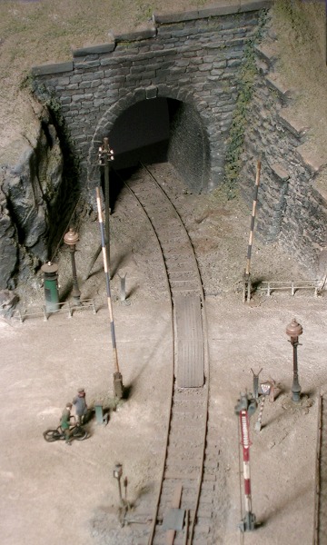 Bahnübergang mit weiß–blauen Schranken und Tunneleinfahrt dahinter, von oben.