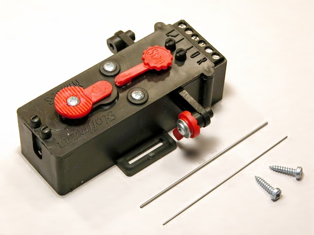 Motorischer Weichenantrieb im schwarzen Kunststoff–Gehäuse, dabei zwei Stelldrähte und zwei Schrauben.