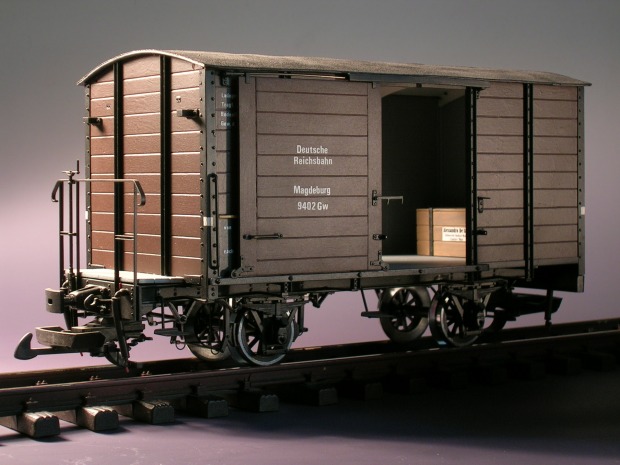 Modellfoto: Brauner, gedeckter Güterwagen mit Bremserbühne und geöffneten Schiebetüren, innen eine Kiste.